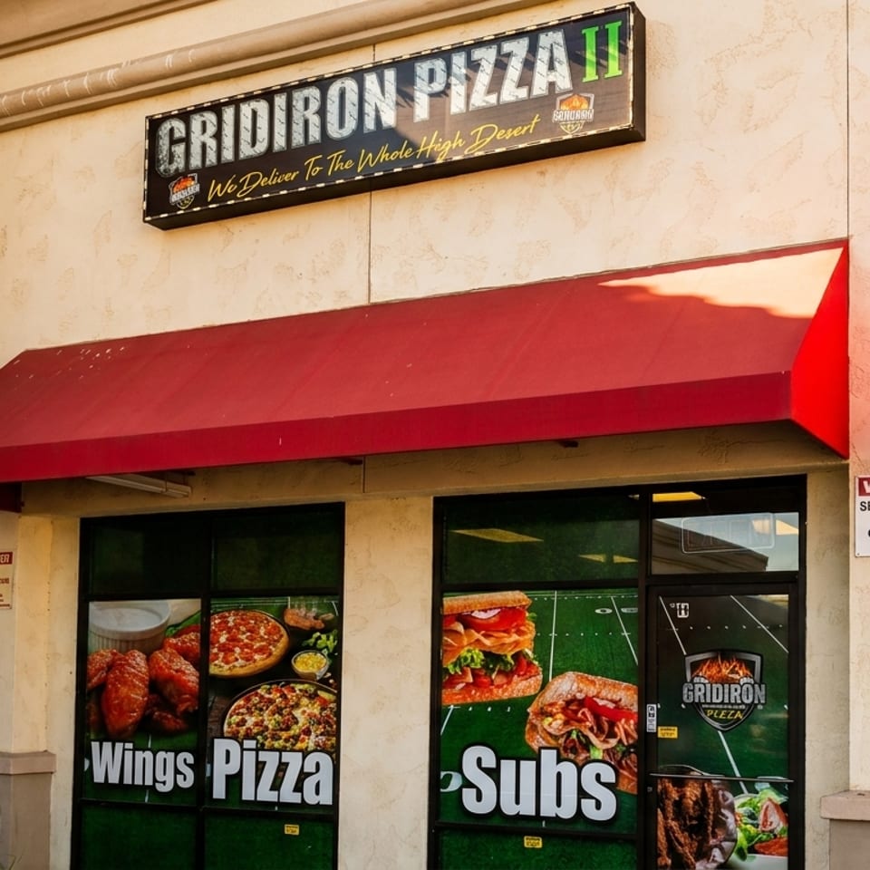 Exterior of Gridiron Pizza II restaurant with a red awning and window decals of pizza, wings, and subs.